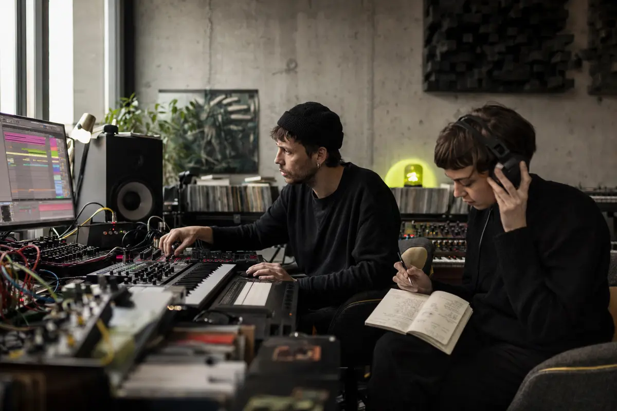 Electronic music producer shaping a track at studio equipment while a collaborator maps the release plan
